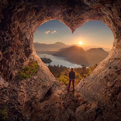 image depicts a figure centered on a heart shaped cave opening looking out on a landscape filled with mountains, forests and lakes. There is a sunrise happening in the distance that gives the image feelings of hope and to be content