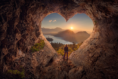 image depicts a figure centered on a heart shaped cave opening looking out on a landscape filled with mountains, forests and lakes. There is a sunrise happening in the distance that gives the image feelings of hope and to be content