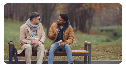 Two dudes sitting on a bench with coffee ups and they chit chat.